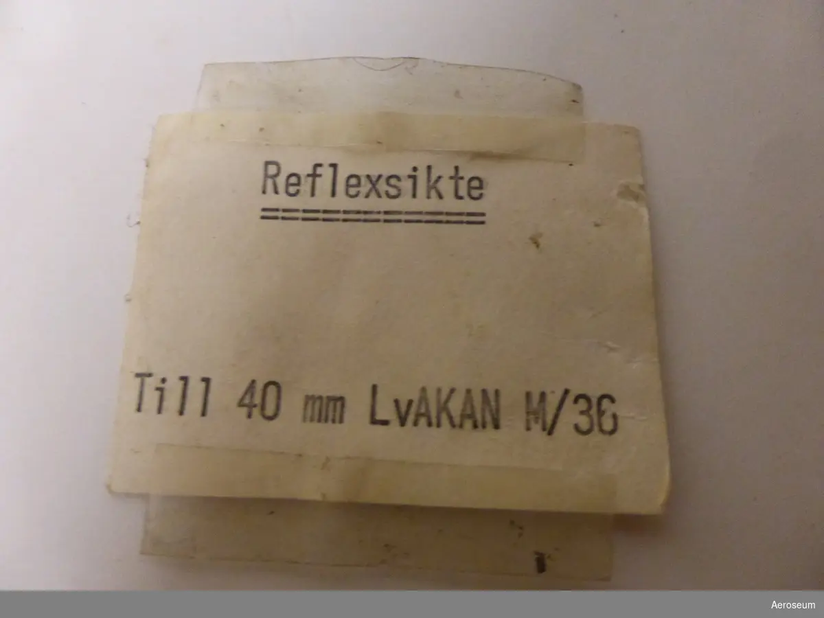 En träfärgad låda med ett reflexsikte i (40 mm Akan M/36). I lådan finns en manual, en pensel, en gul imskyddsduk ("Den svenska imskyddsduken"), en sladd, två bitar tjockt glas i i varsin storlek (den mindre är mörkfärgad), en vit tejprulle, två extra små glödlampor, en gulnad mätskiva med siffror på.

Utöver det hänger en etikett på lådan som verkar handla om olika åtgärder och när de gjorts, med viss text som inte går att tyda. Åtgärder eller kontroller verkar varit utförda 24/10 1962, 1/10 1963, 18/8 1966, och 22/4 (?) 1968. På ettiketten är det stämplat "Instrumentservicegruppen FO 32/31 - OBK V. Frölunda 5". På andra sidan står det att ettiketten är en kontrollapp för tygmateriel. Det går också att läsa "Mtrlslag: Reflexsikte 40 mm Akan", "Nr 1957 80", "Fel, erforderliga åtgärder, brist etc klass 3: ua [?]", och "Tillhör 42801"

På manualens framsida står det "AGA-BALTIC REFLEXSIKTE"
