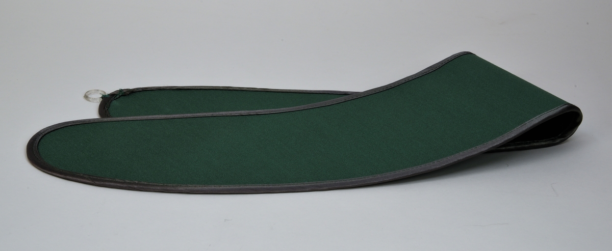 Ett gardinomtag av grönt siden, med kant av grönt/silverskimrande tyg. På omtaget sitter det två fastsydda plastringar som fästs ihop med en metallhyska, även den fastsydd.