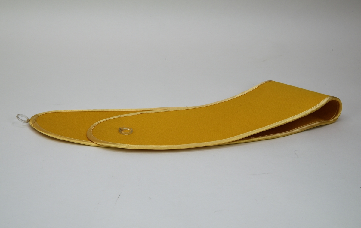 Ett gardinomtag av gult siden, med kant av gultonat skimrande tyg. På omtaget sitter det två fastsydda plastringar som fästs ihop med en metallhyska, även den fastsydd.