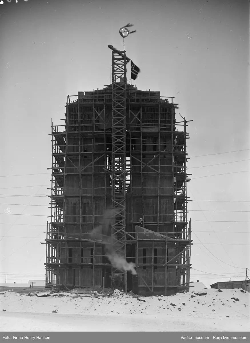 Vadsø 19.5.1956, kirka er under bygging. På toppen av kirketårnet er det er norsk flagg og noe som antagelig er en mønsåskrans. Dette er vanlig å henge opp på nybygg når taket er tett.