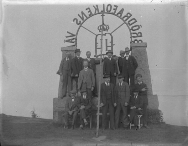 En grupp män på baksidan av kung Oscar II:s monument på Tabergs topp i Småland. Kungens valspråk "Brödrafolkets väl" bildar en båge mellan kraftiga naturstenspelare på ett fundament. Männen har klivit upp på monumentet och några sitter på bänkar nedanför. 

Monumentet tillkom 1895 i samband med att Oskar II invigde järnvägen mellan Nässjö-Jönköping, via Taberg, anlagd av Halmstad-Nässjö Järnvägs AB (HNJ). Efter att ha skrivit sitt namn på en berghäll nära stationen for konungen upp till bergets topp och därefter till Norrahammars bruk. Där spelade brukets musikkår folksången medan brukspatron J W Spånberg eskorterade majestätet in till gjuteriet där hans namnchiffer omgifvet av ett "Brödrafolkens väl" göts i sanden. HNJ lät så småningom utforma detta monument med det gjutna namnchiffret.
(Se även bild HB658)