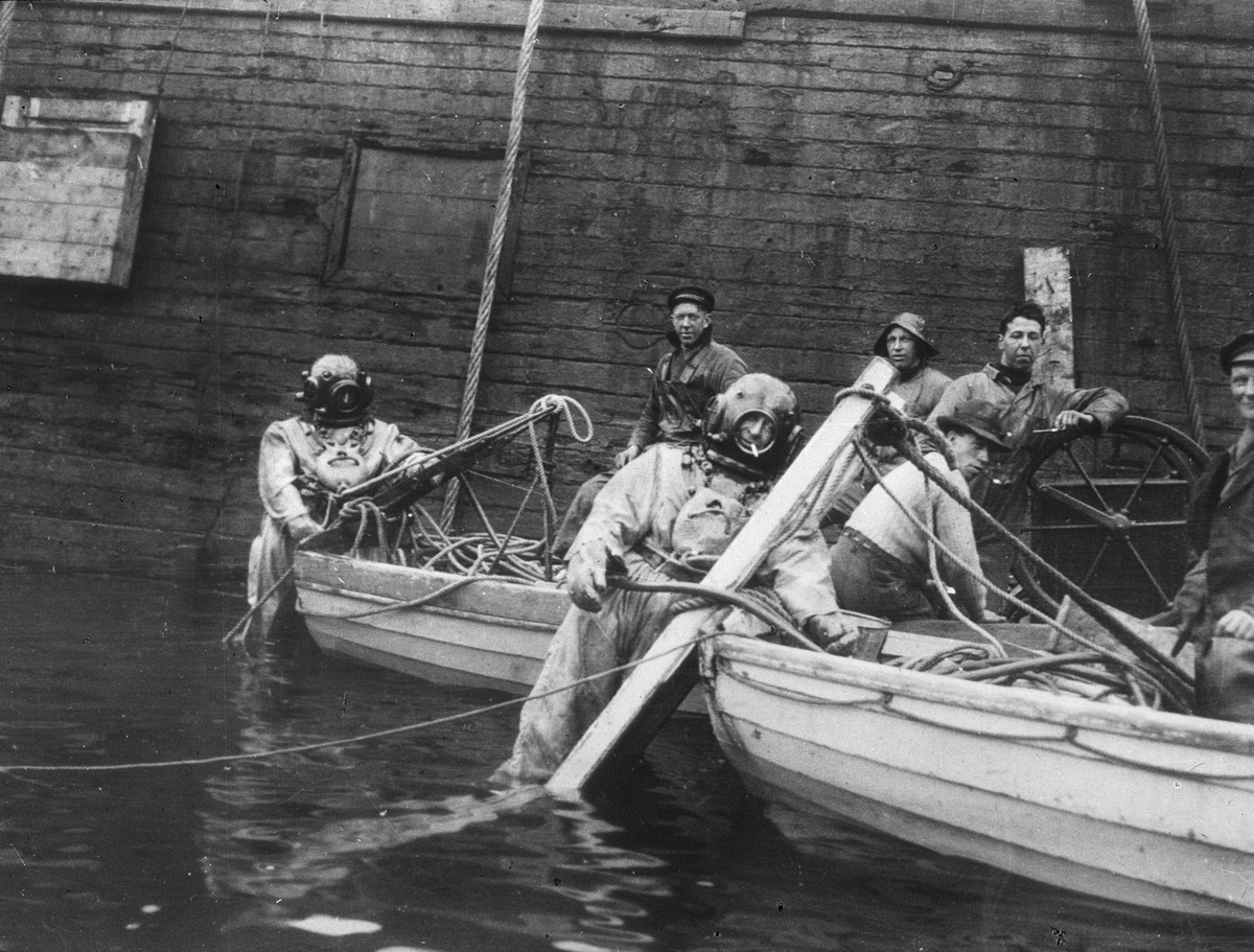 To hjelmdykkere og fem arbeidsfolk ombord i to lettbåter. Båtene ligger inntil en skuteside. Hjelmdykkeren med sigarett i munnen er Bernhard Meier Sebak (1894-1976).