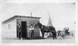 To menn og hest med vogn foran en hytte. En hund ligger ved 