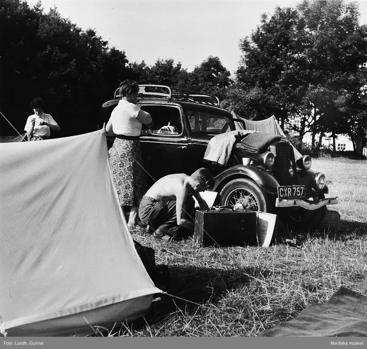 Två personer med packning vid en engelskregistrerad Ford Y årsmodell 1934. I förgrunden ett tält.