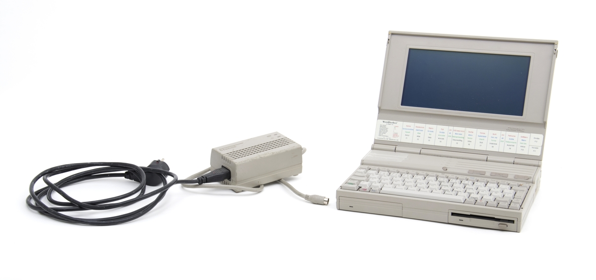 En lys grå PC med lader. Datamaskinen er en av de eldste bærebare PC i denne størrelsen og en av de først med både innebygd harddisk og en diskettstastjon. Batteristasjonen er reparert med tape. PC er ellers i god stand og virker fortsatt.
