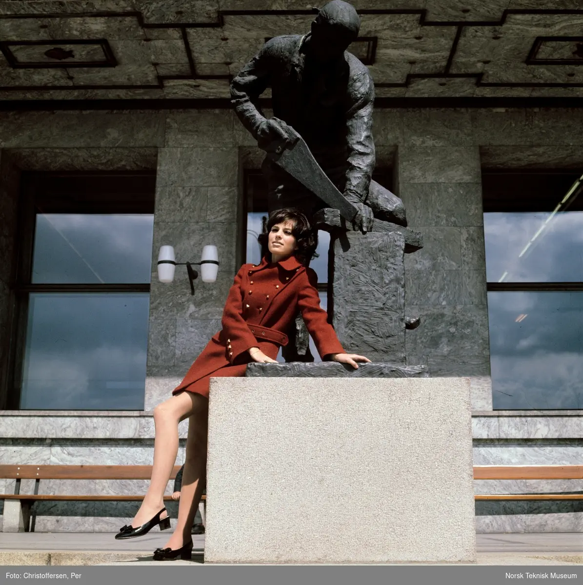 Motebilde i farger. Kvinnelig modell i rød ullkåpe, sittende på statuen "Snekker" (1954) av Per Palle Storm foran Rådhuset i Oslo, høsten 1968. Bildet er en del av en serie der noen er publisert i ukebladet Det Nye samme høst.