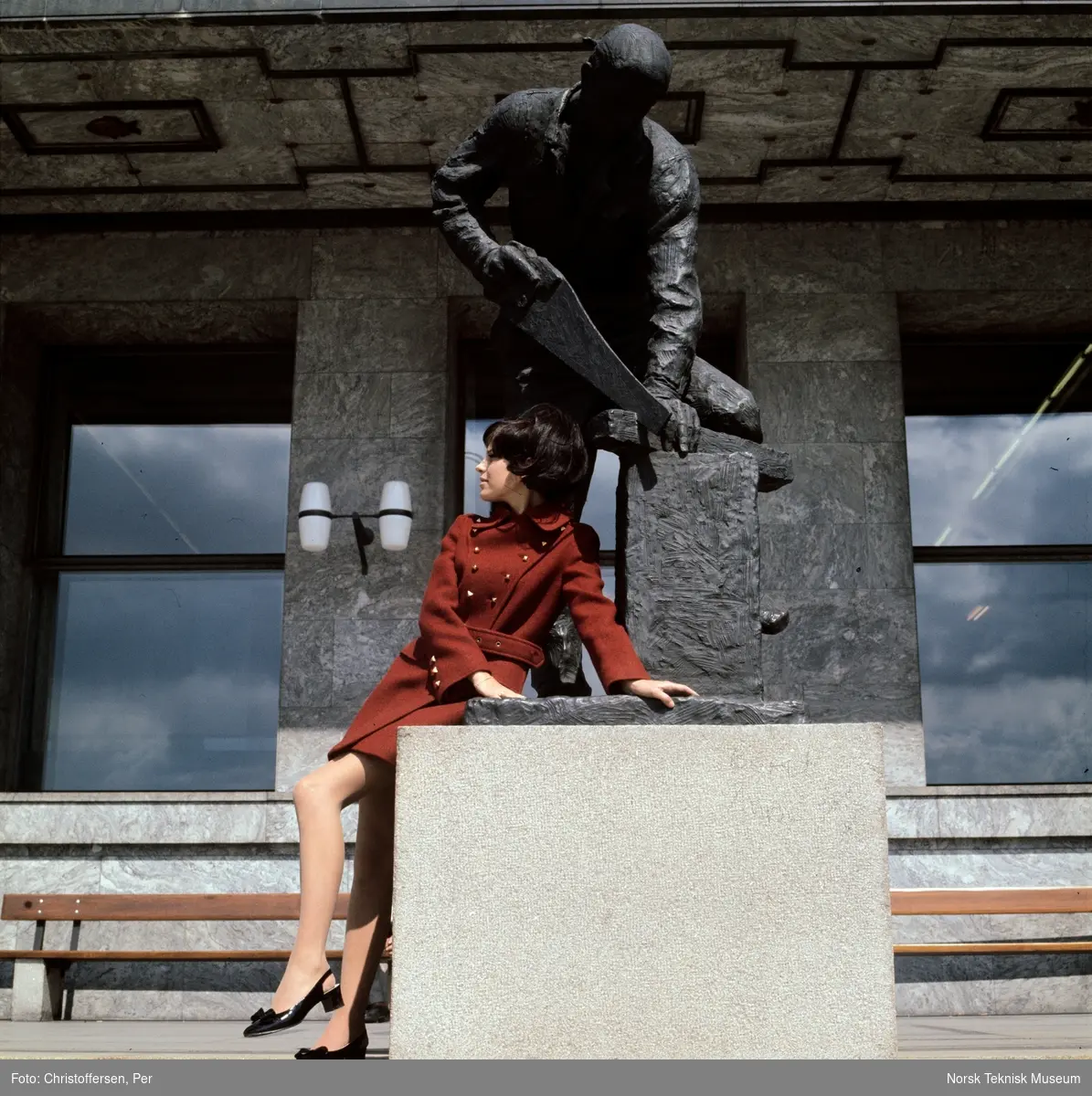 Motebilde i farger. Kvinnelig modell i rød ullkåpe, sittende på statuen "Snekker" (1954) av Per Palle Storm foran Rådhuset i Oslo, høsten 1968. Bildet er en del av en serie der noen er publisert i ukebladet Det Nye samme høst.