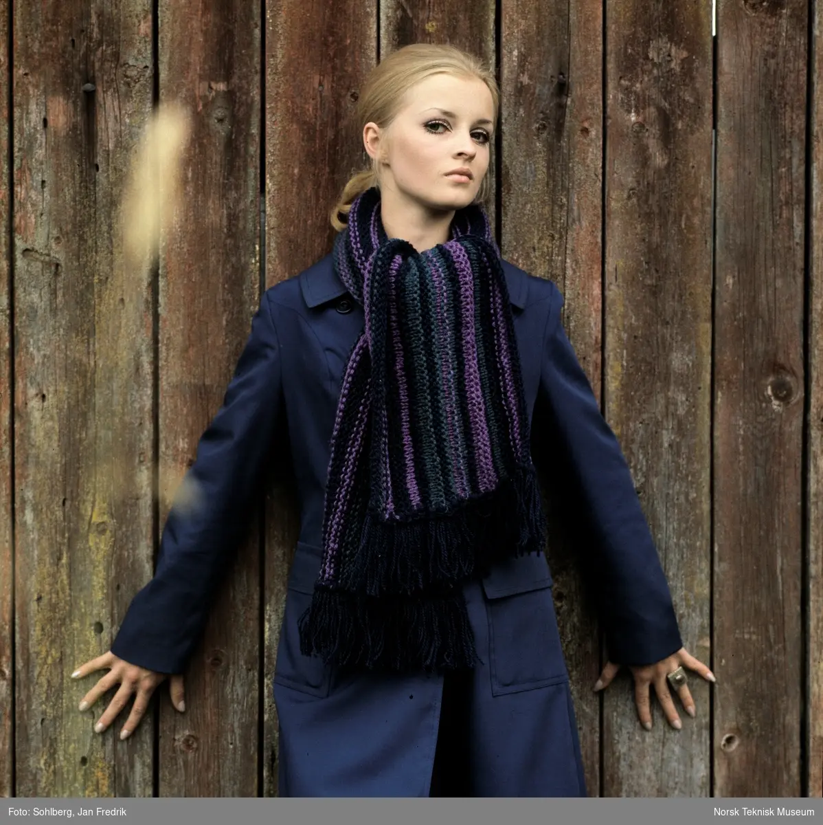 Tidlig norsk motefotografi. Kvinnelig modell står ved plankevegg. Hun har mørk lilla kåpe og stort strikket skjerf.