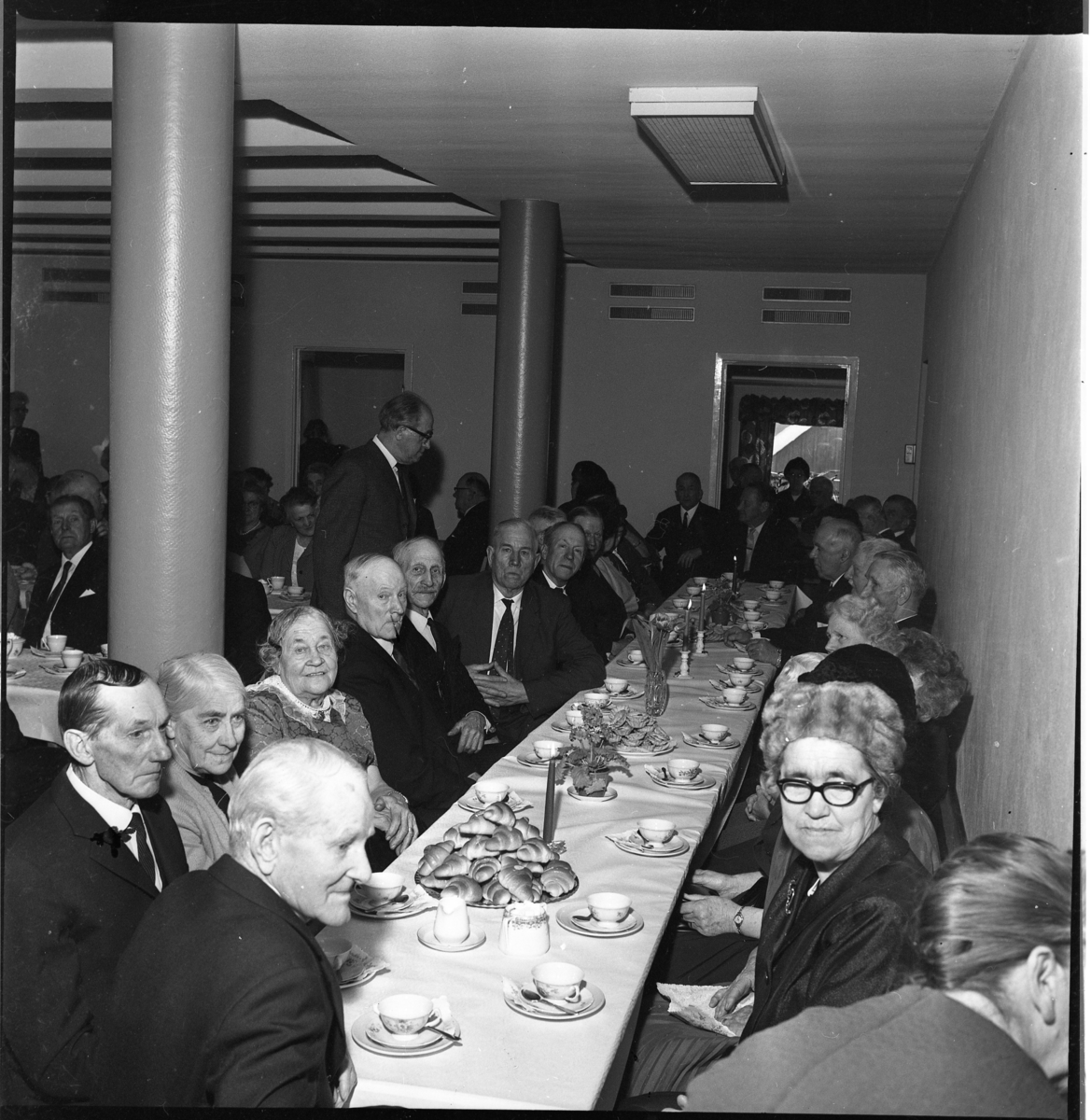 En samling äldre män och kvinnor sitter vid dukade kaffebord, troligen i Filadelfiakyrkan i Gränna. Märtha Persson sitter som trea på vänstra sidan och tittar mot fotografen.