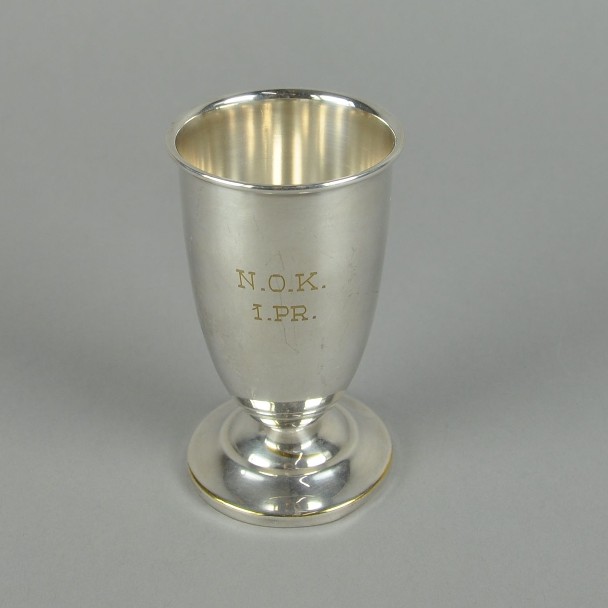 Pokal av metall, muligens sølvplett. Sylindrisk form med innsnevring nederst mot utgående sokkel. Pokalen har profilert dekor på sokkel.
