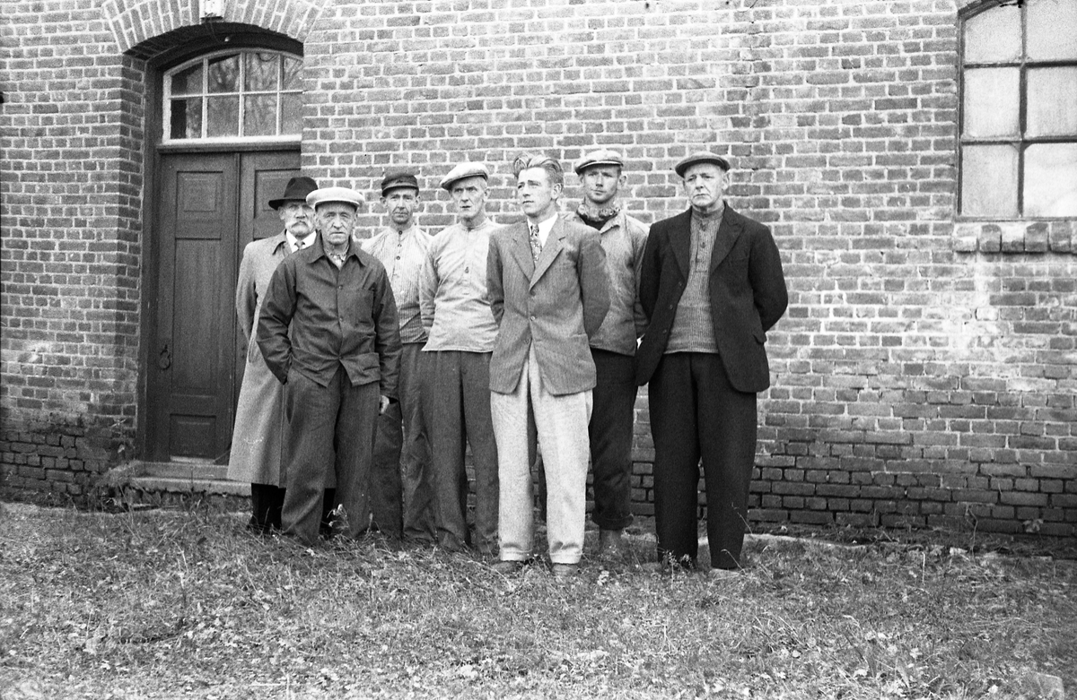 Sju uidentifiserte menn avbildet foran en teglsteinsvegg. Bildet, som er fra høsten 1956, kan muligens være fra potetmelfabrikken/brenneriet på Lena.