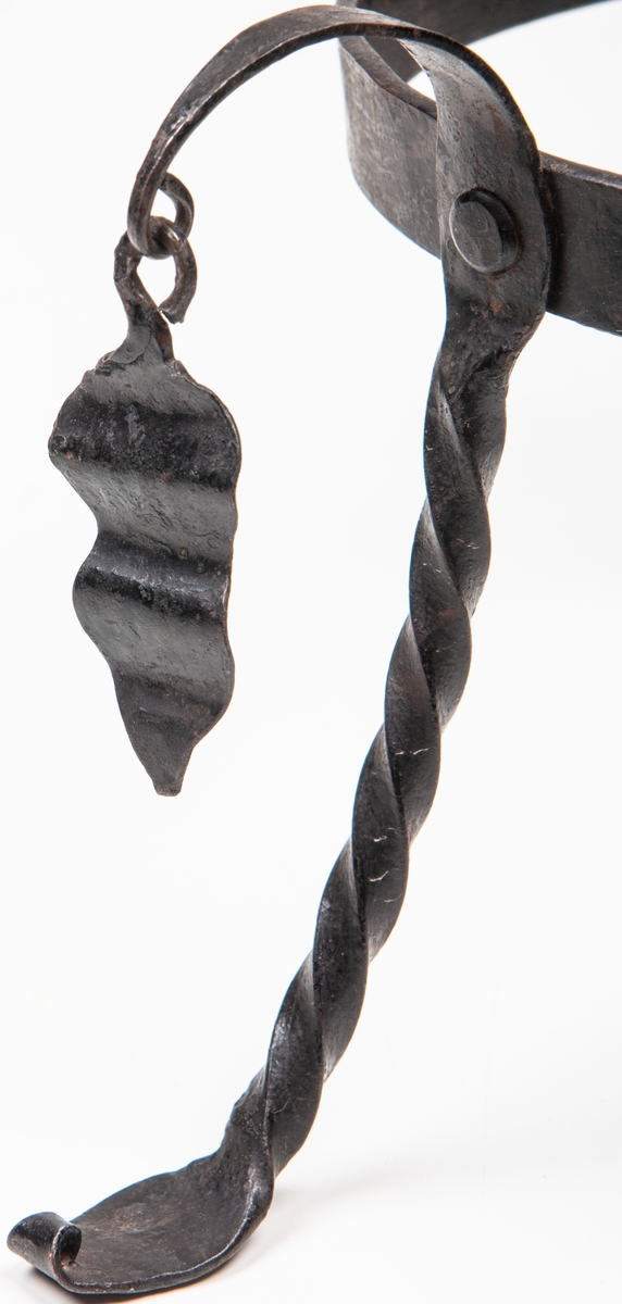 Trefot av järn. Smidd. Spiralvridna ben och vidhängande hängen.
Höjd 11,5 cm. Diameter 15,5 cm.