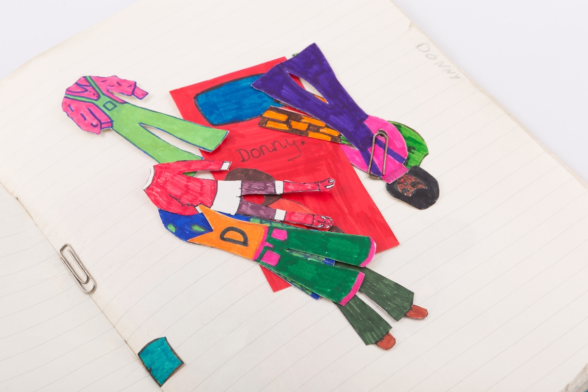 Hjemmelagde papirdukker oppbevart i ei mappe laget av ei  gammel skolebok med plasttrekk. Hver dukke ligger i sin lomme merket med navn.