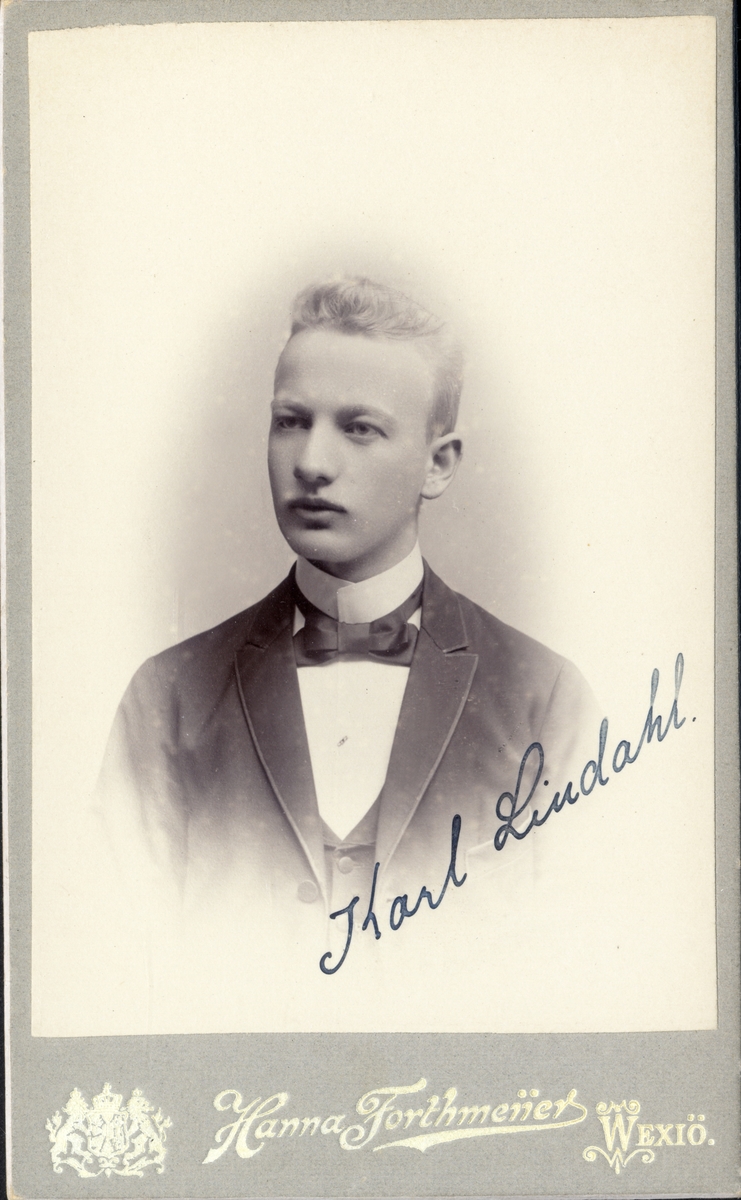 Foto av en ung man i kostym med väst och stärkkrage med fluga. På snedden i nedre högra hörnet finns ett namn skrivet:
"Karl Lindahl". 
Bröstbild, halvprofil. Ateljéfoto.
