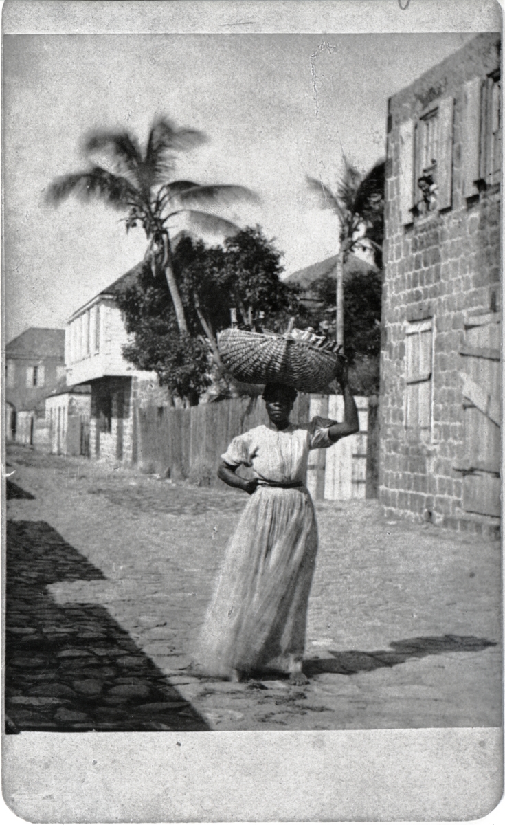 Gatubild från Gustavia. 
Kvinna med korg på huvudet. Ett barn tittar ut från ett fönster.