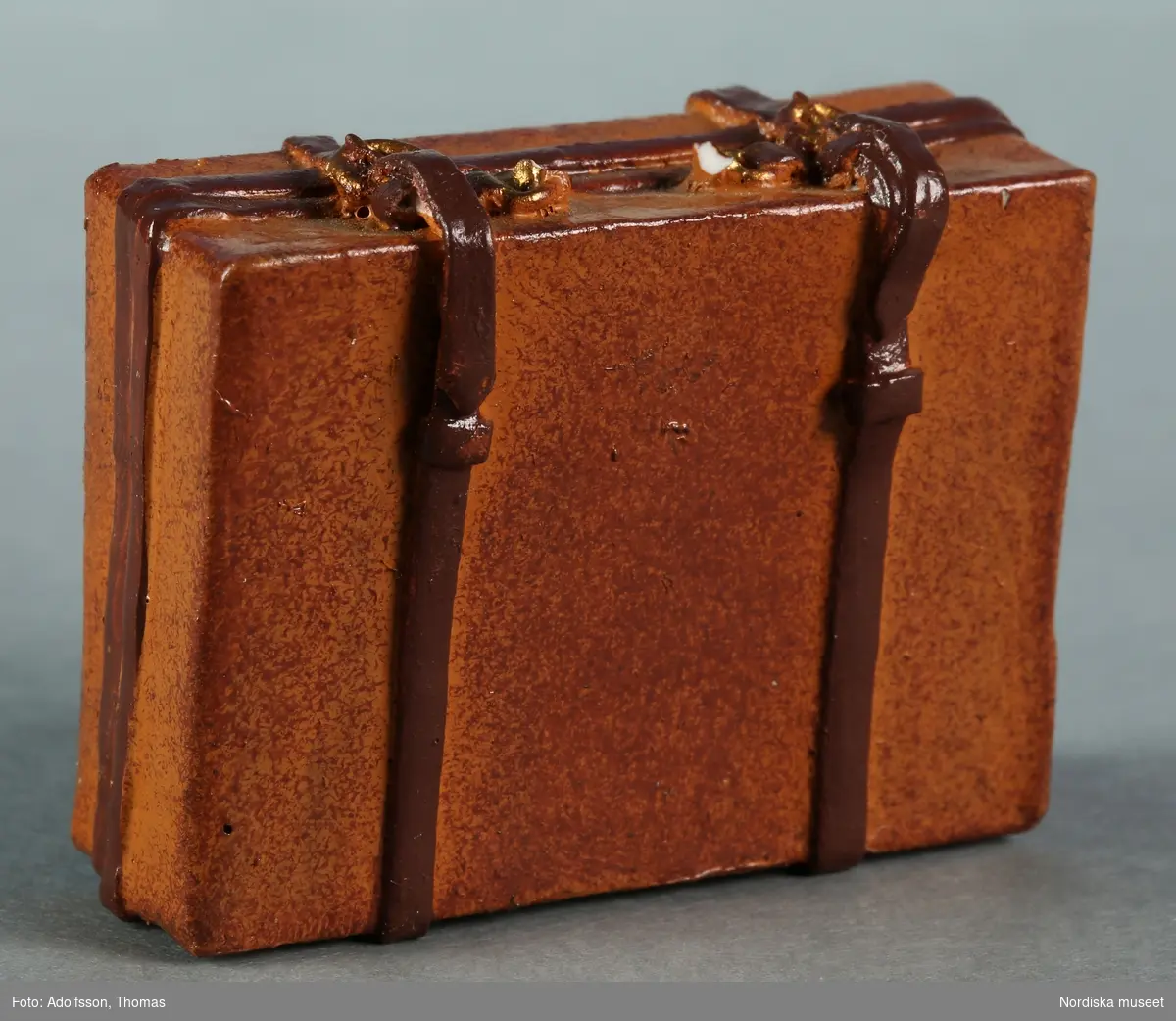Resväska tillverkad av gips eller konstmassa och målad i två olika bruna nyanser. Ska stå i dockskåpets vindsutrymme.