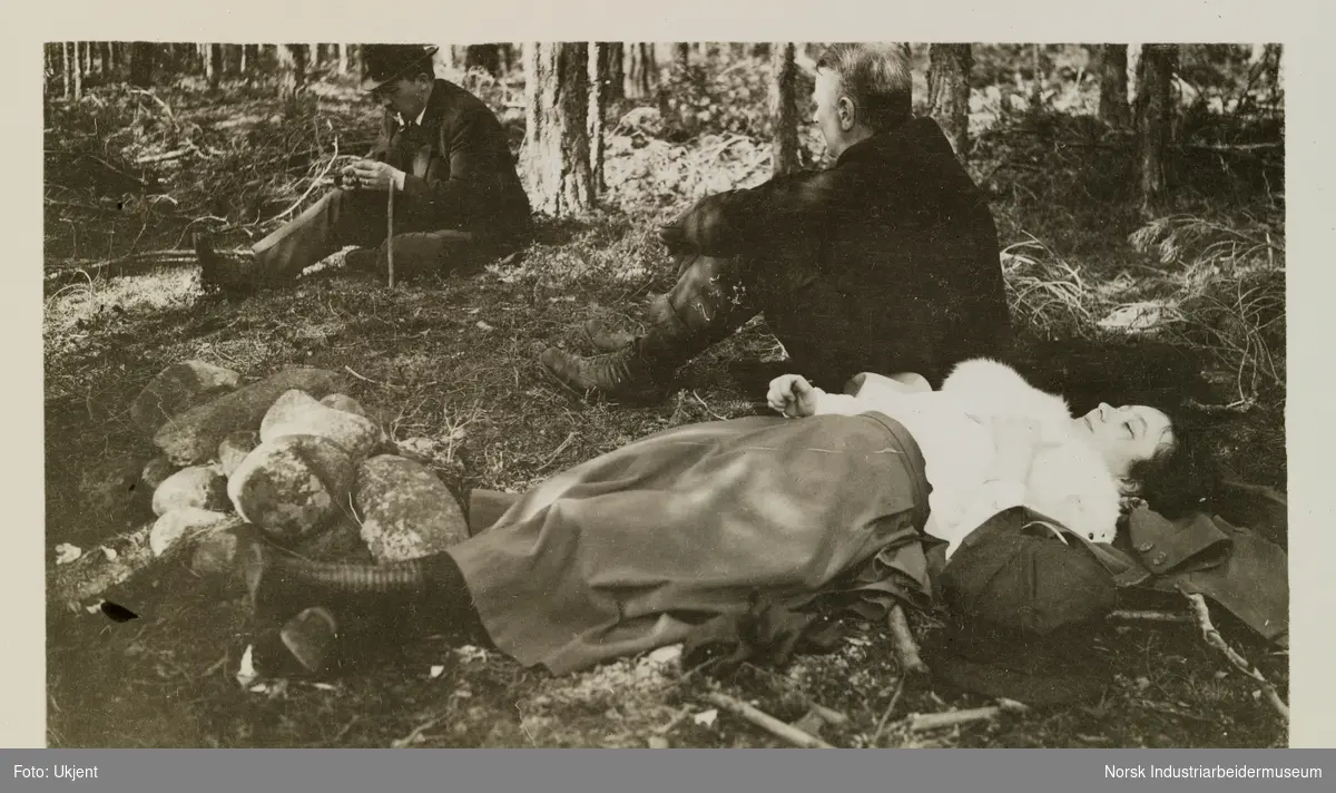 Kvinne ligger på rygg og to menn sitter, ute i naturen