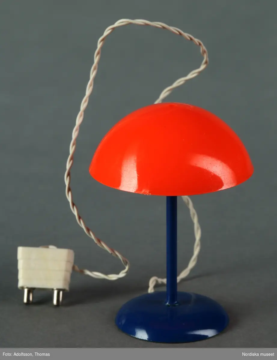 En a) golvlampa och b-c) två bordslampor av plast, metall och syntettyg, samtliga med elektriska kablar och små glödlampor. Golvlampan har röd skärm medan borslamporna har orange respektive gul skärm. Hör till dockskåpets pojkrum, andra våningen. Den minsta gula lampan ska stå på sekretären (NM.0333264 a-d) medan de andra står på golvet.