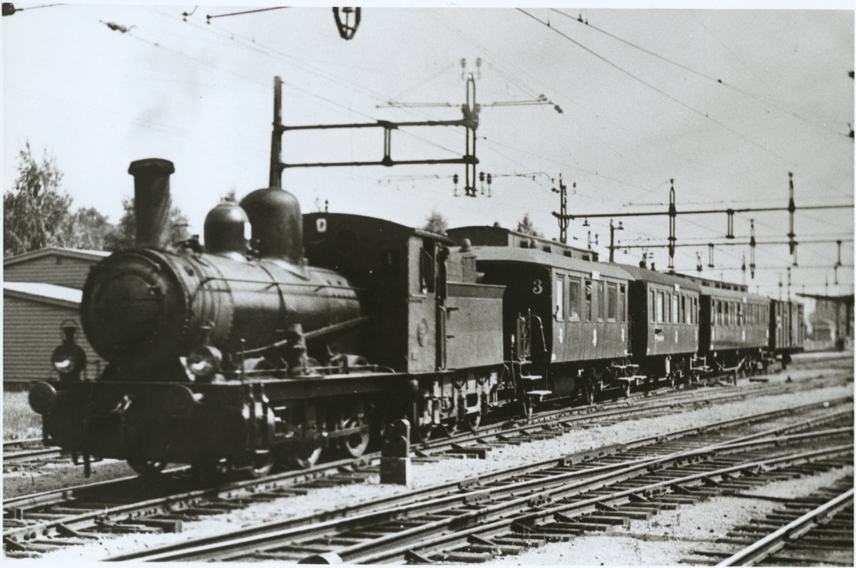 Tåg med ånglok och personvagnar på elektrifierad bangård.