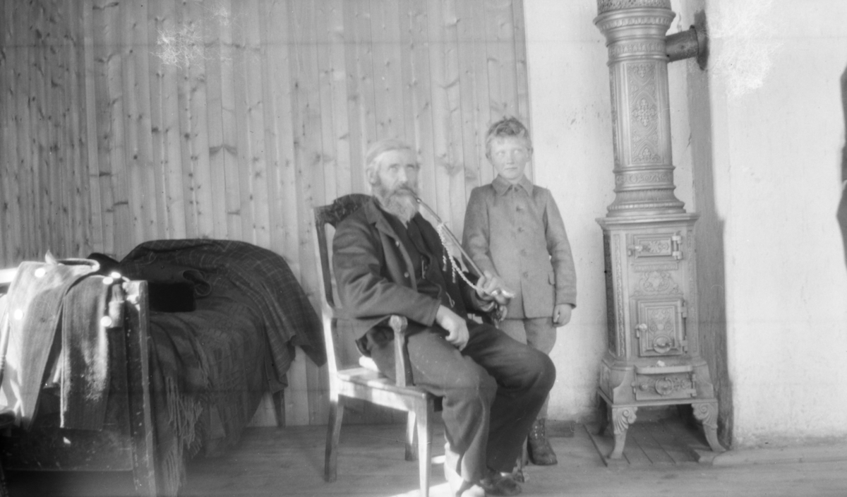 Eldre mann med krokpipe og gutt, innen ved en rund - muligens på Linjordet i Østre Gausdal