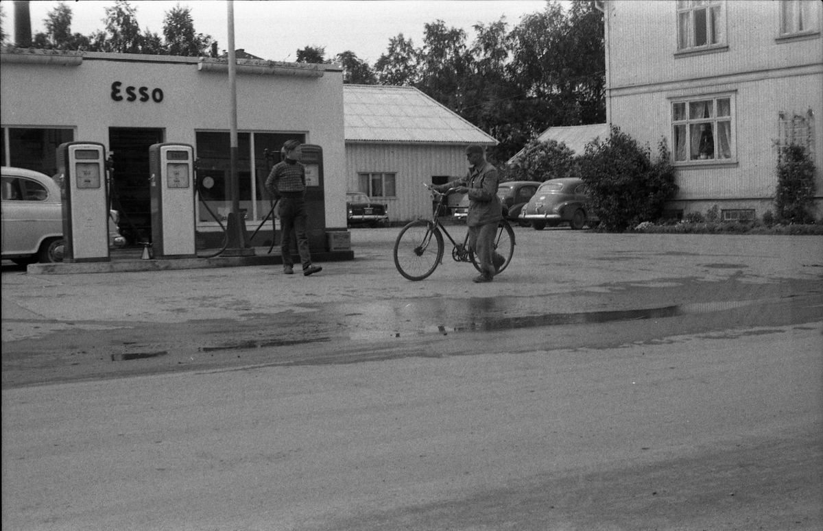 Esso bensinstasjon på Lena april 1958. En mann triller en sykkel forbi, en annen står foran pumpene, og henvender seg kanskje til han med sykkelen. Personene er ikke identifisert. Bildet er tatt gjennom vinduet i Lena Foto & Radio i "Raubua".
