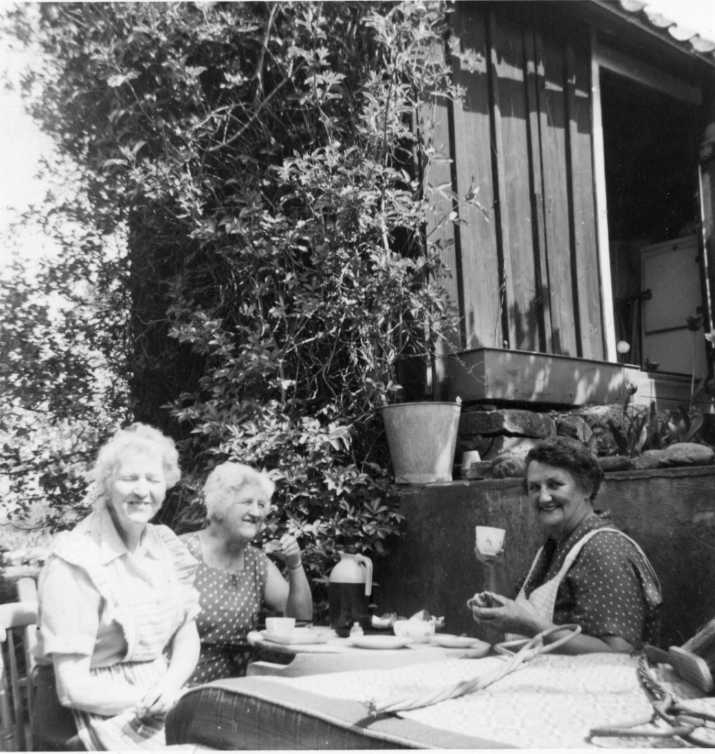 Tre kvinnor tar en kaffepaus i hushållsarbete. De sitter runt ett bord med termos och kaffekoppar. Intill ligger en matta och mattpiskare. Kvinnorna är Lisa och Märta Lindholmer samt Sigrid Johansson till höger.
