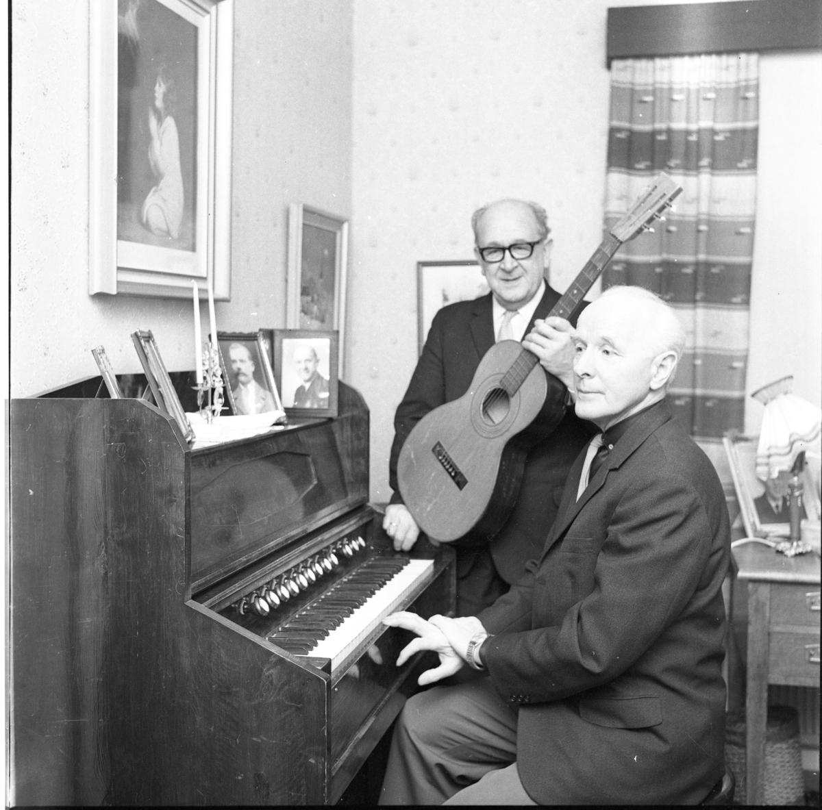 Carl Öst i glasögon står med en gitarr intill Martin Persson som sitter vid en orgel. På orgeln står en ljusstake med två ljus samt ramade porträttfotografier av män.