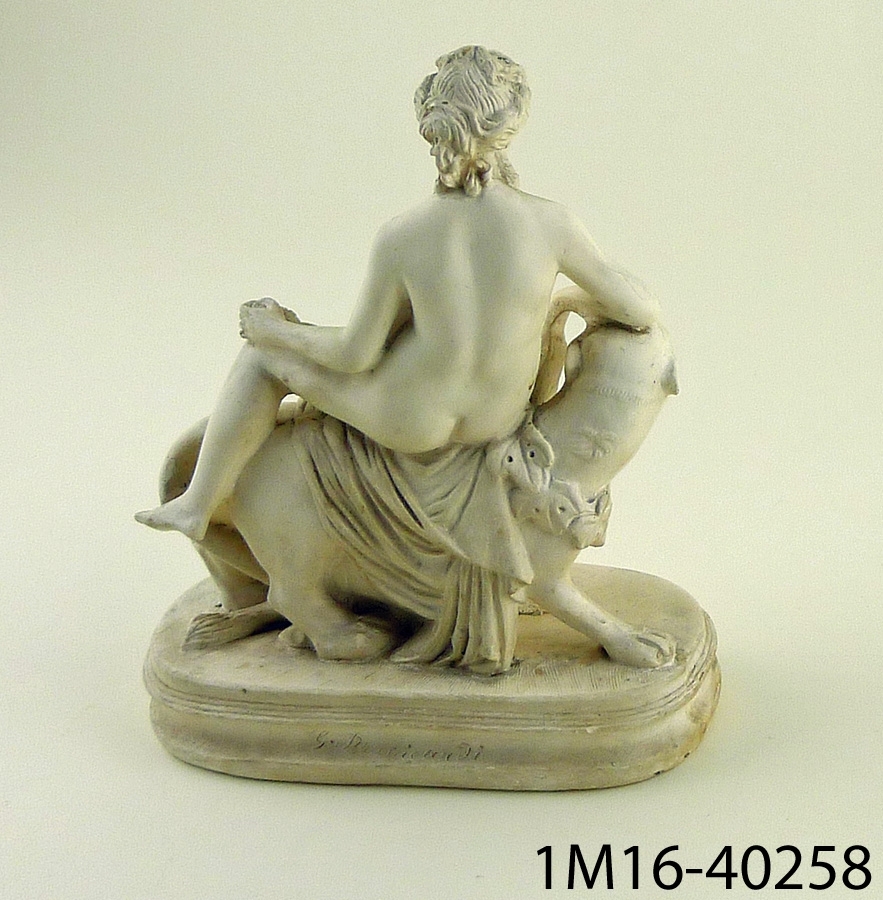 Gipsskulptur föreställande naken kvinna sittande på lejoninna. Kvinnan sitter på ett tygstycke och håller en vindruvsklase i sin ena hand.
