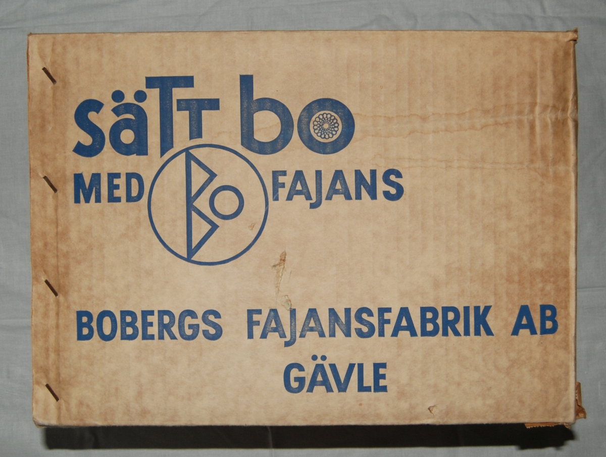 Låda av wellpapp. Tillverkad för Bo Fajans i Gävle med text på ena långsidan: Sätt bo med Bo Fajans - Bobergs Fajansfabrik Gävle. På mostående sida en bild av keramikföremål.