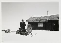 James Coward står i snøen ved snøskuter og hytte med torvtak
