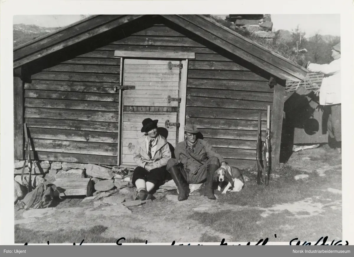 Aslaug, Hans Erik og hund sitter langs hyttevegg. Jaktgevær er stilt opp langs hyttevegg. Til Høyre sees mann ved torvtak