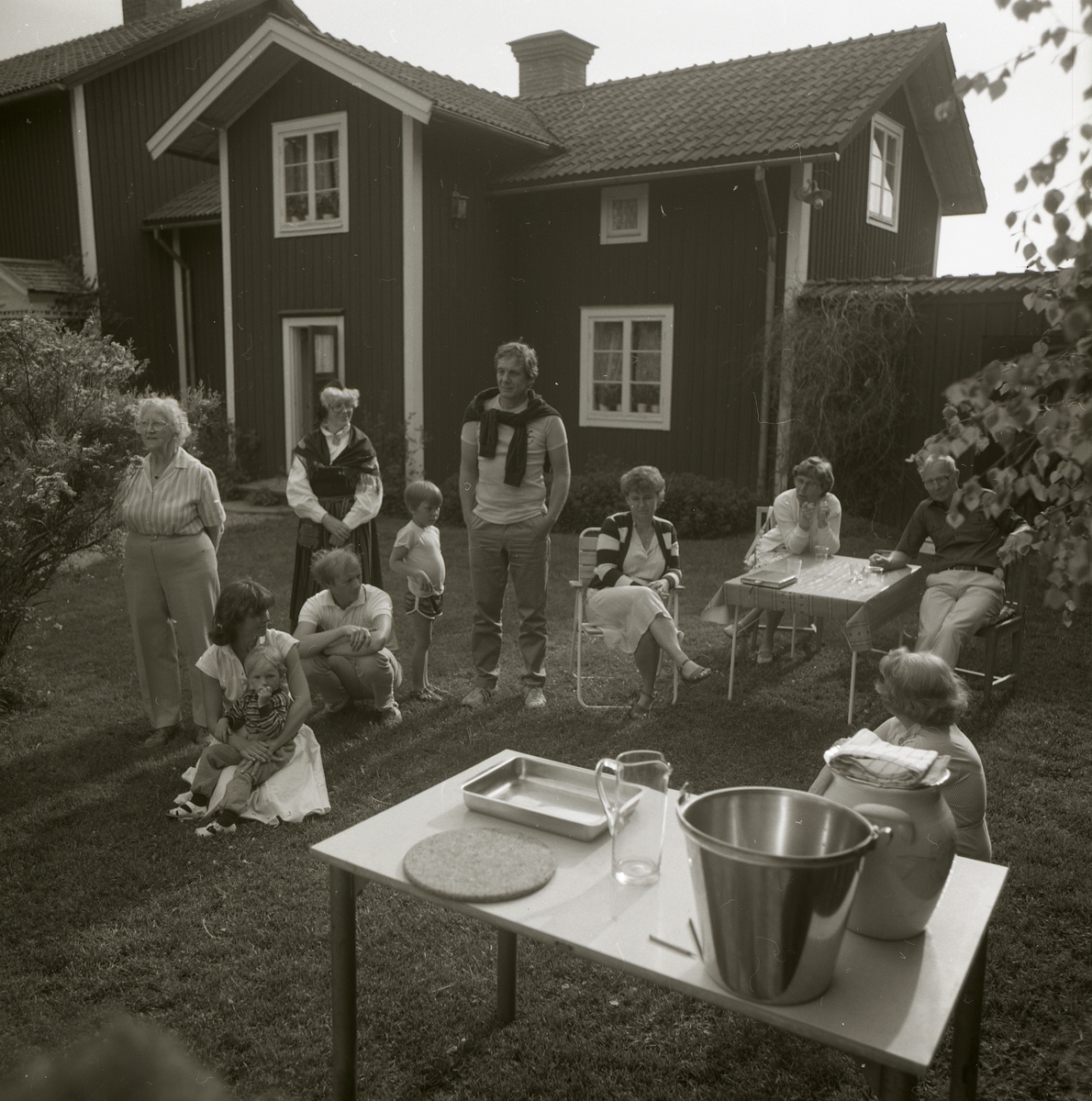 Några människor står samlade på gårdsplanen vid gården Sunnanåker under ett midsommarfirande, 22 juni 1985. Några människor sitter vid bord.