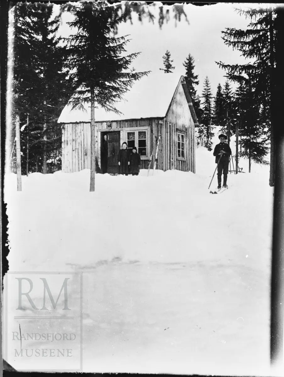 Et lite hus i skogkanten. Ei dame titter ut i dørgløtten. To unger står i snøen utenfor. En gutt på ski til høyre i bildet.