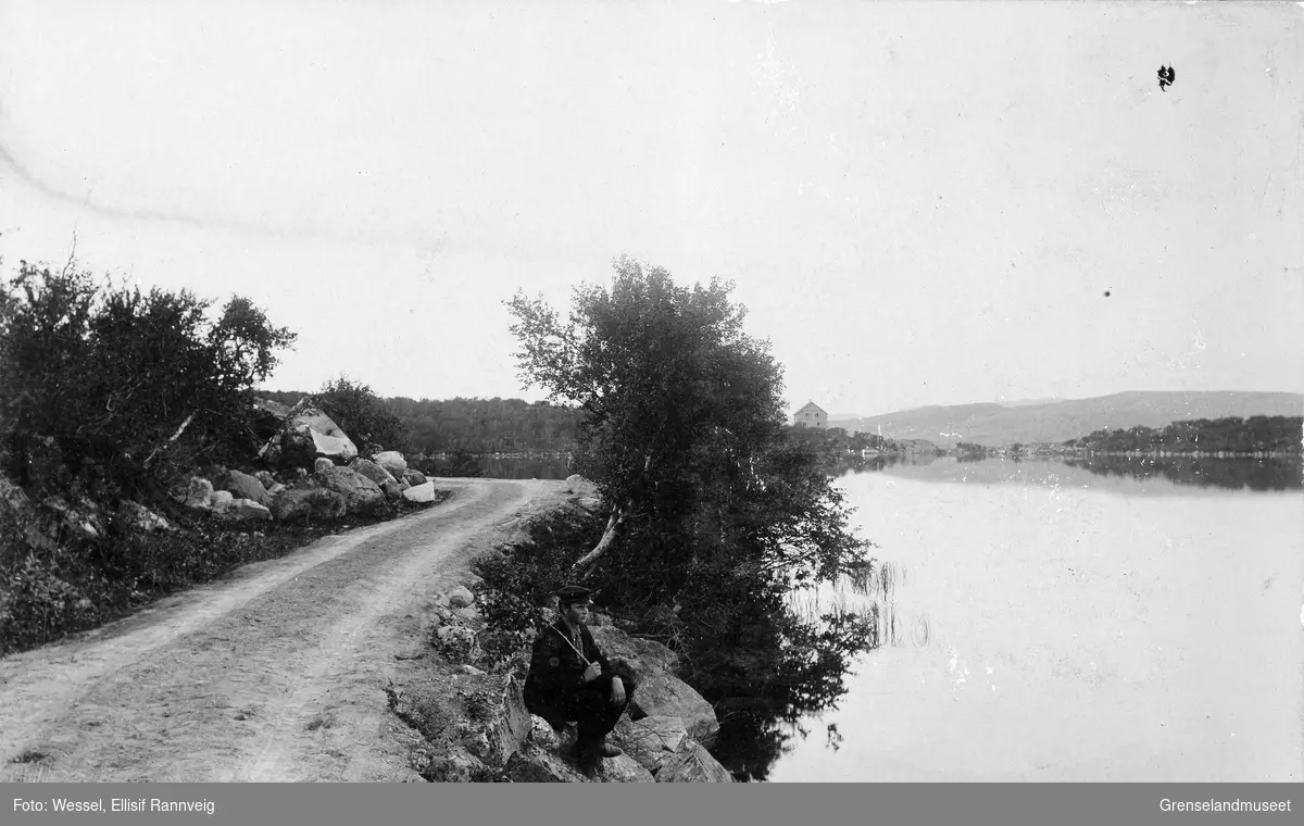 Veien inn til Kirkenes sett mot nord. Førstevannet (Første Kirkenesvand) på høyre side. En ung mann sitter i veikanten og ser utover vannet. Et hus skimtes i bakgrunnen.