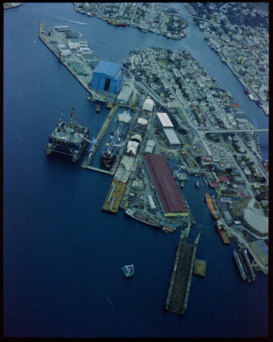 Flyfoto av Haugesund Mekaniske Verksted med omegn. Et stort skip ligger i tørrdokken, riggen "Ocean Liberator" ligger rett utenfor og en rigg ligger ved Storasundskjer. Nordsjøhallen er ferdig og tatt i bruk.