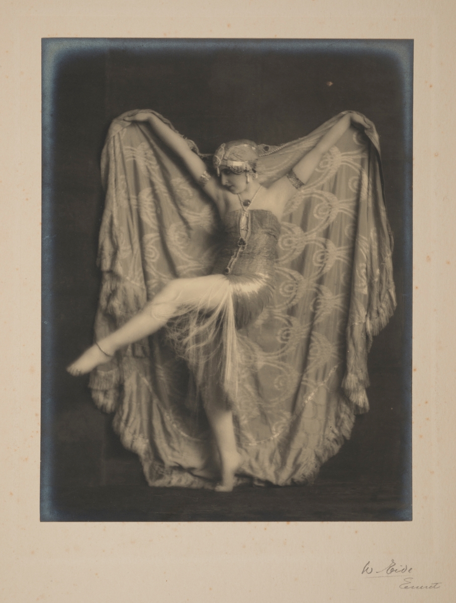 Kunstnerisk portrett av den berømte russiske ballettdanseren Vera Fokina, som holder et stort mønstrete teppe bak seg.
Fra 'Salome - De syv slørs dans'.