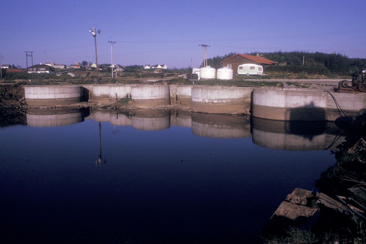 En klynge med betomgkummer, plassert i kanten av en stor dam. I bakgrunnen står en del utstyr og en del bebyggelse.