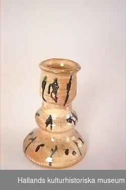 Ljusstake av bränd keramik. Handdrejad. Glaserad, melerad i gul-beige med dekorationer av svarta och gröna streck och prickar. Profilerad med utsvängd fot, i botten märkt H Cederholm, Varberg, 1972.  Övre diameter:60 mm. 