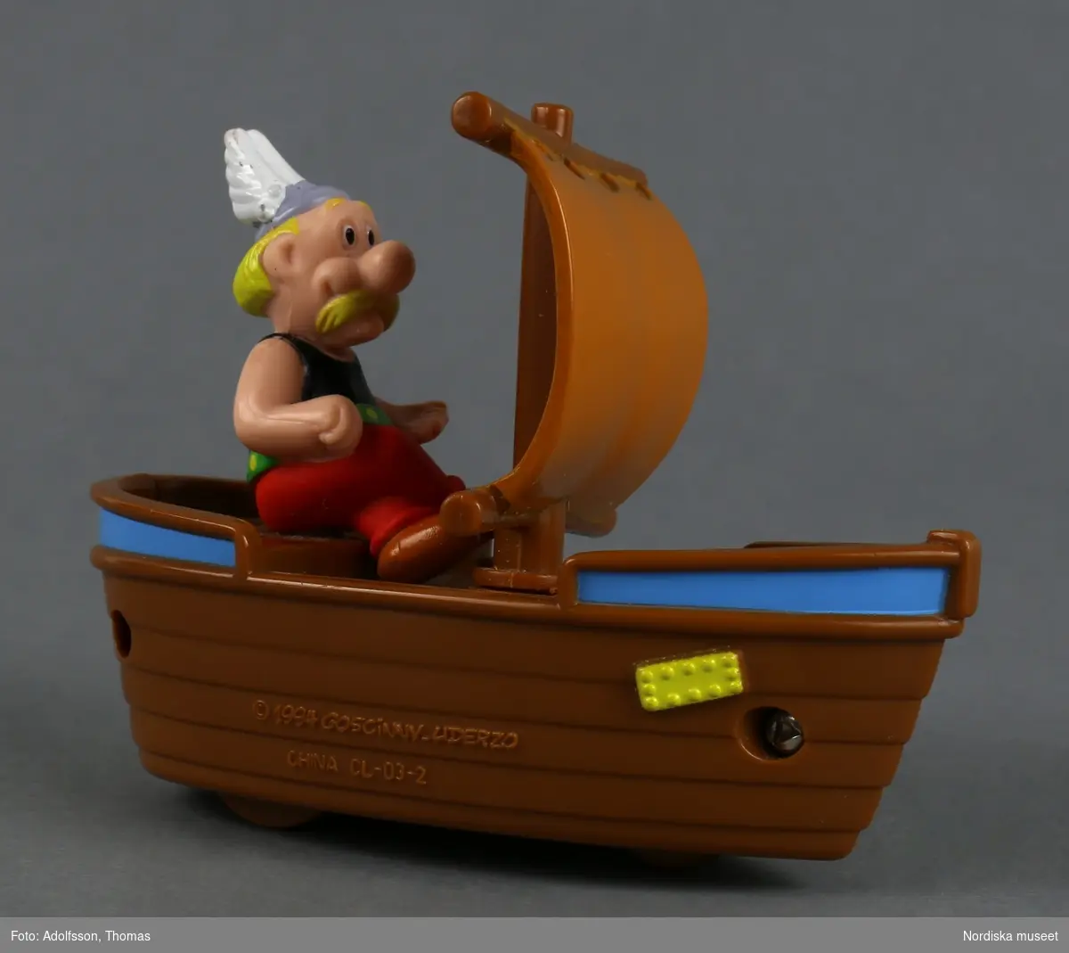 Leksaksbåt av brun plast med figuren Asterix. I mitten av båten ett segel och på undersidan tre små hjul. Om man drar båten mot underlaget rör sig seglet fram och åter.