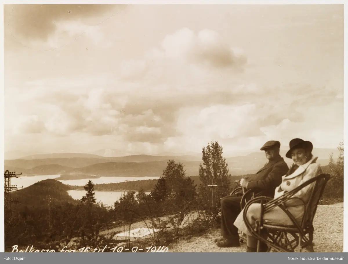 Mann og kvinne med hatt og jakke sitter i stoler og nyter utsikten ved Bolkesjø hotell