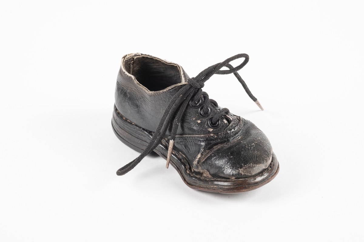 Et randsydd barnesko (venstre sko) av lær. Skoen har snøring med runde lisser. Maljene er av metall. Sålen er av lær og er forsterket med spiker på hælen og ved tuppen på undersiden.