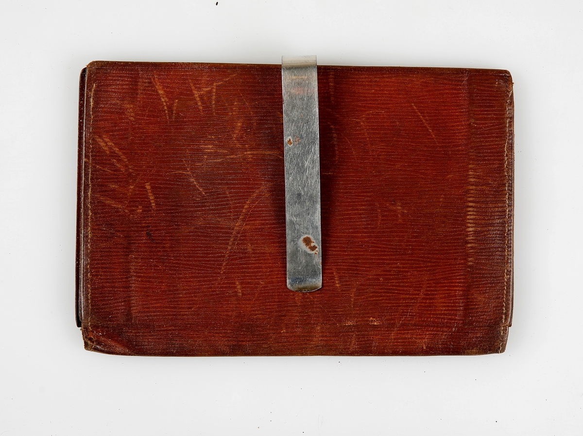 Stor lommebok som holdes sammen med en metallklemme. Inni er det to trekkspillommer og en lomme lukket med reim gjennom hempe. 
