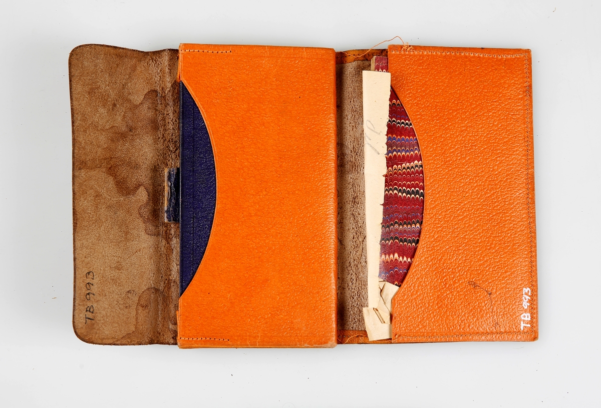 Avlang lommebok uten lukning. Bak er det en hempe som antyder at det har vært en reim til å lukke lommeboka med. Inni er det tre trekkspilllommer og en stikklomme. I stikklommen ligger det en notisbok. Lære inni er farget oransje og blått. 