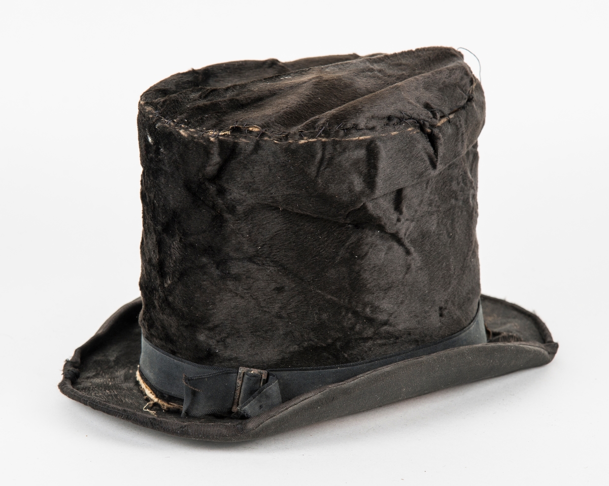 Hatt, høg svart flosshatt i silkefloss. Ripsband med ei spenne rundt pullen, kanta med ripsband rundt. Fóra inni med lerret. Saumen i pullen har gått opp,