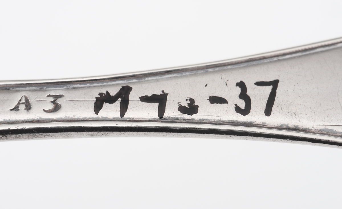 Sked av silver.
Svensk trubbig modell. Skaftets baksida har ingraverade initialer; "JBK, MCU". Stämplarna sitter längre ner på baksidan. 

Inskrivet i huvudbok 1984.