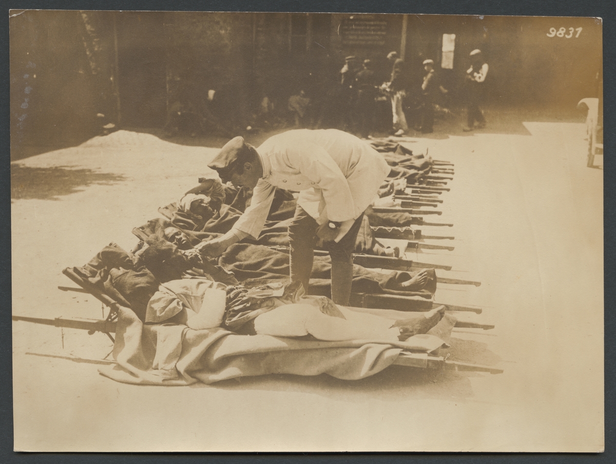 Bilden visar en rad sårande afrikanska soldater som ligger på bår på marken. En man i vit rock och uniformsmössa delar ut cigaretter.