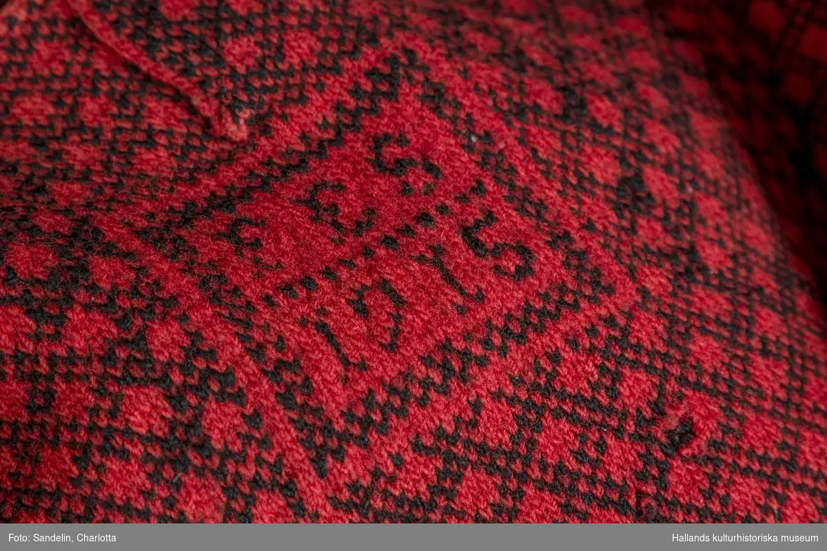 Bingetröja stickat av rött yllegarn med mönster i svart garn. I halsen 1 knapp, därunder "FES 1915". Hål vid höger ärmhåla.