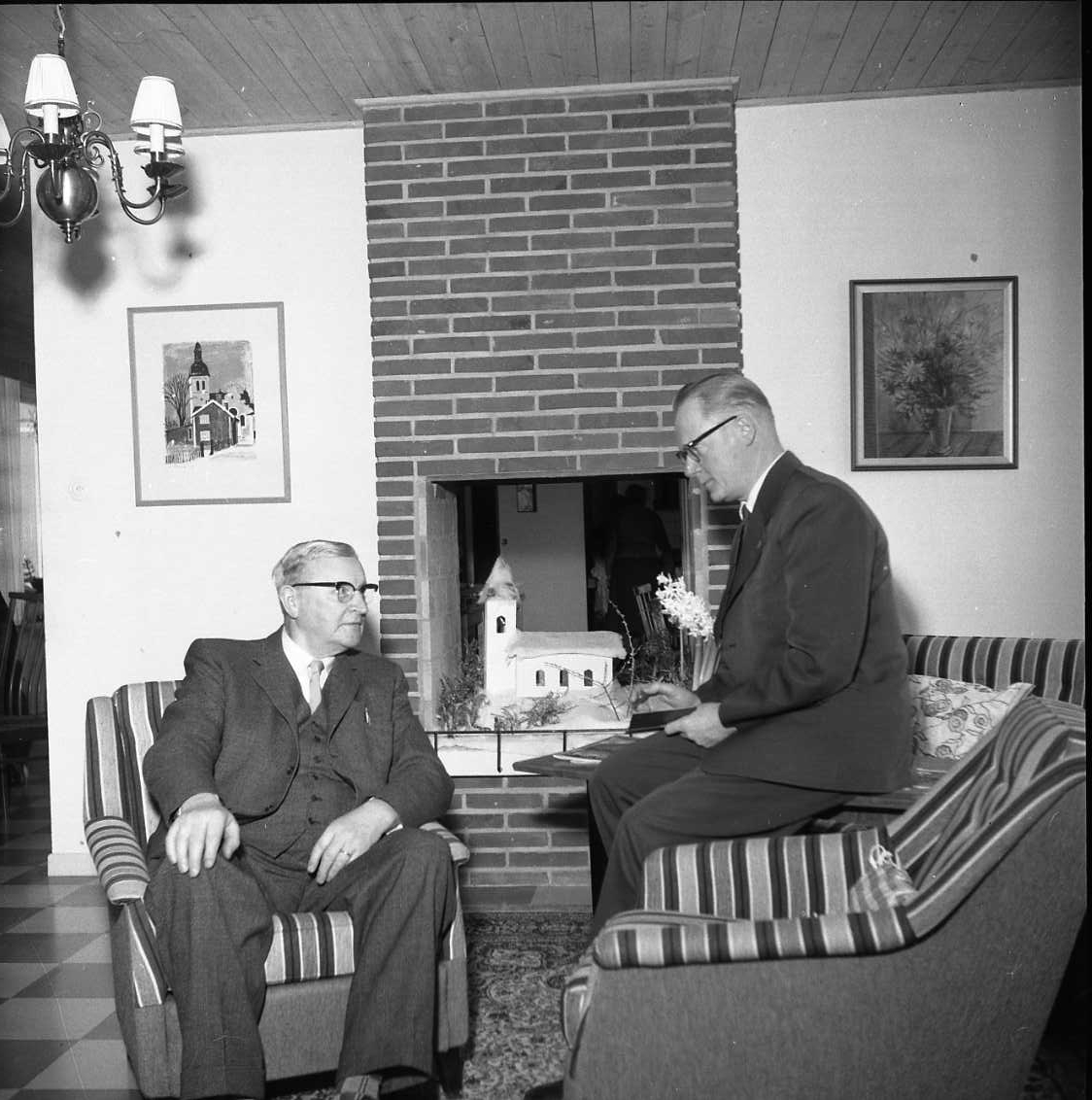 Interiör från en hemmiljö. Gustaf Rahm sitter i en fåtölj och samtalar med Percy Heldebrandt som sitter på ett bord.