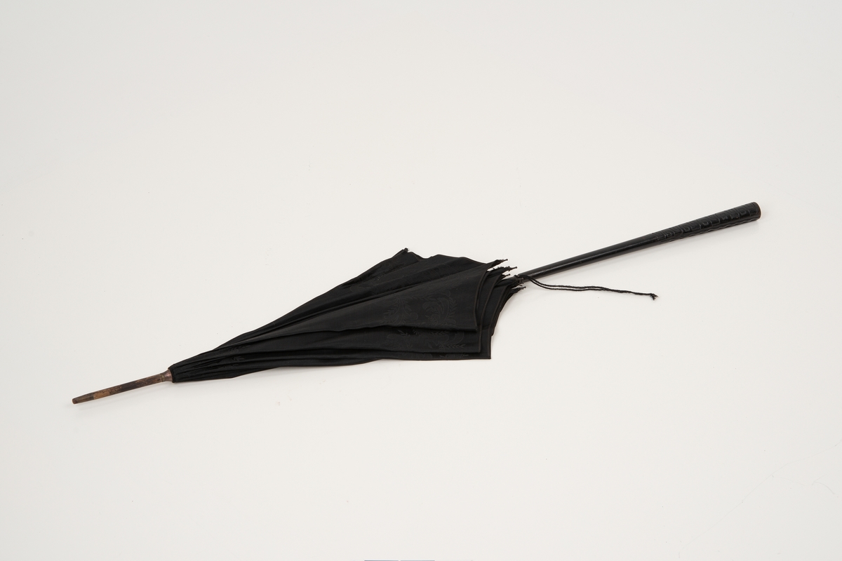 Svart parasoll med treskaft. Utskjæringer nederst i skaftet. Åtte metallspiler, trukket i svart silkedamask med organisk mønster.