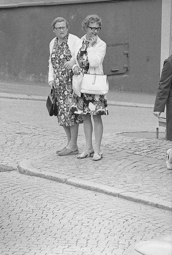 Två kvinnor med glasögon,  i mönstrad klänning och ljus kofta med handväska på armen står vid ett gatuhörn och tittar åt sidan. De står troligen vid hörnet Brahegatan och Ribbagårdsgränd.